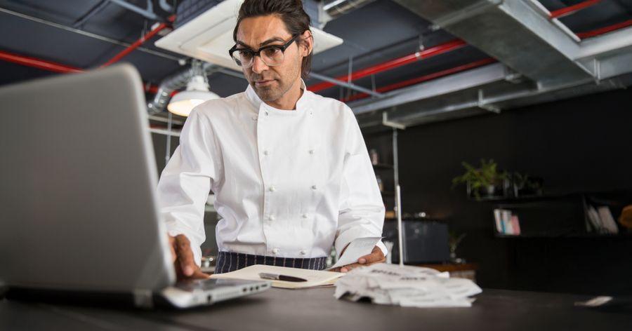 Chef in a white uniform and glasses working on a laptop in an industrial-style kitchen, with papers and a pen nearby.