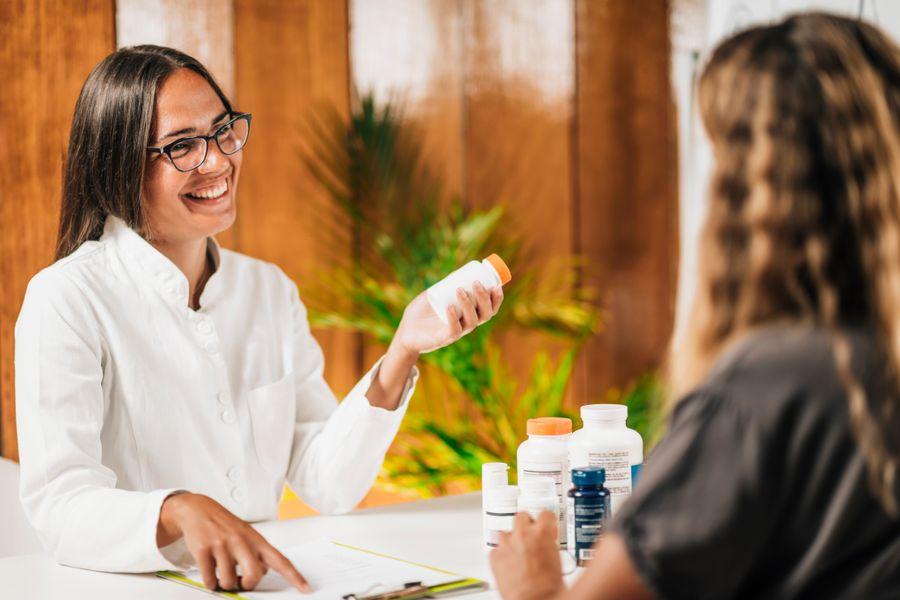 A smiling pharmacist in a white coat discusses medication options with a customer, holding a pill bottle. Various bottles are on the table.