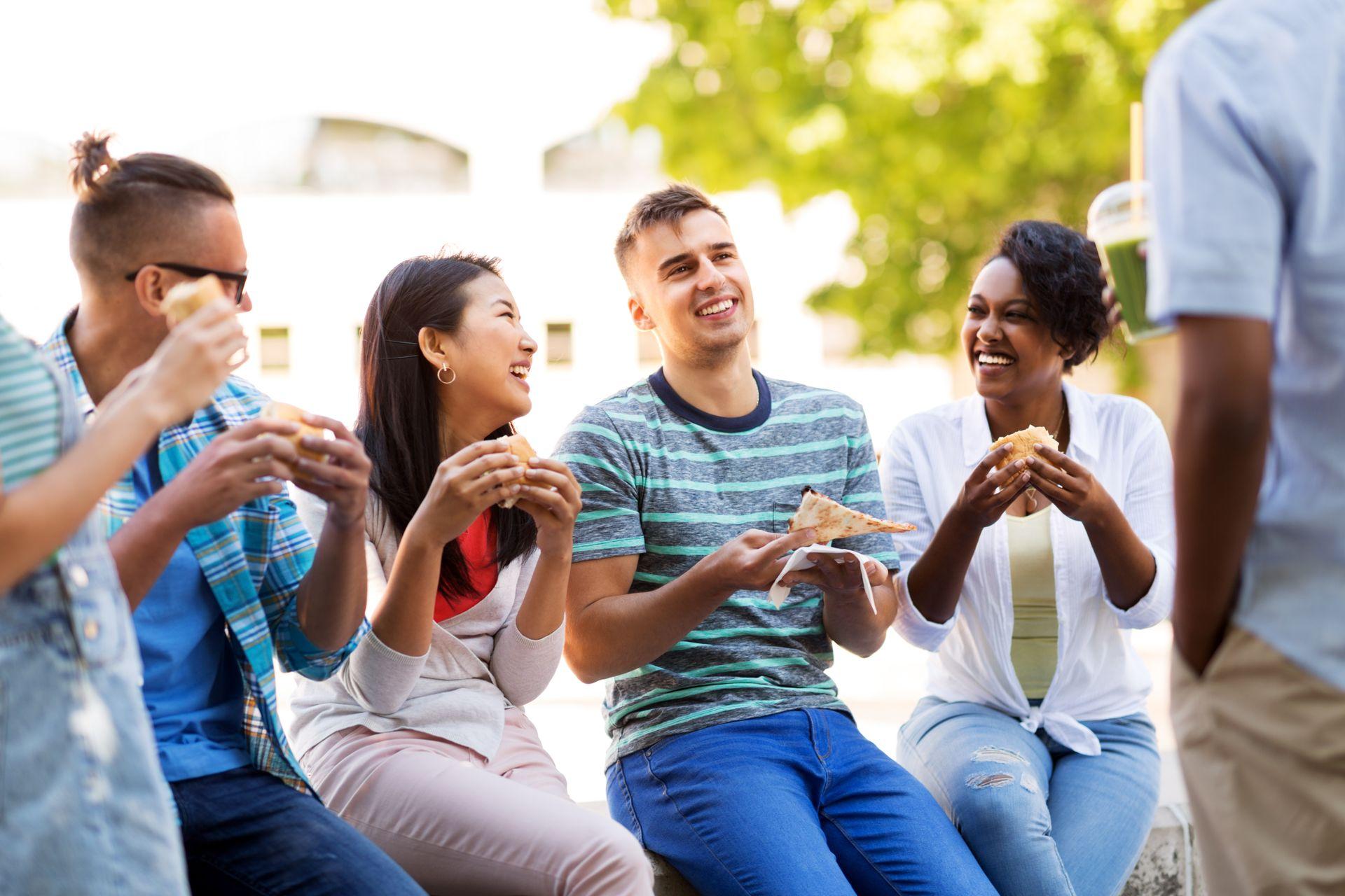 A group of five people sitting outdoors, smiling and eating pizza together on a sunny day.